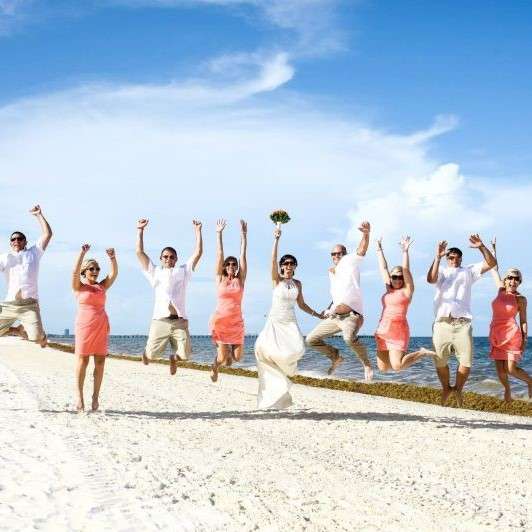 bridesmaids in pink sundresses and groomsmen in white shirts and tan pants jump in a horizontal line