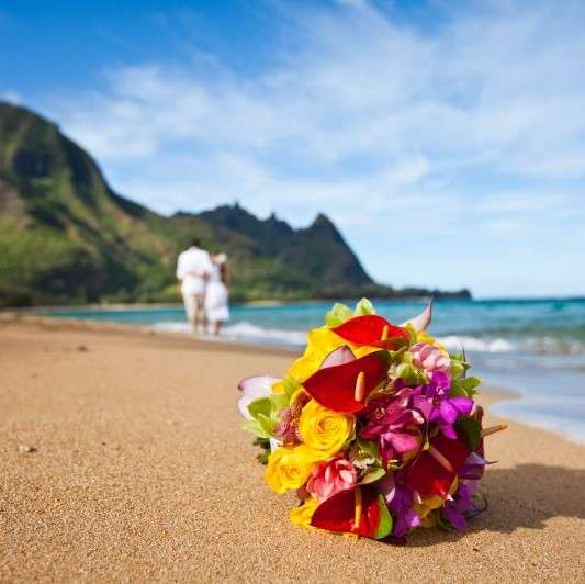 bride's bouquet flowers in red, purple, yellow on a beach with hawaii green cliffs